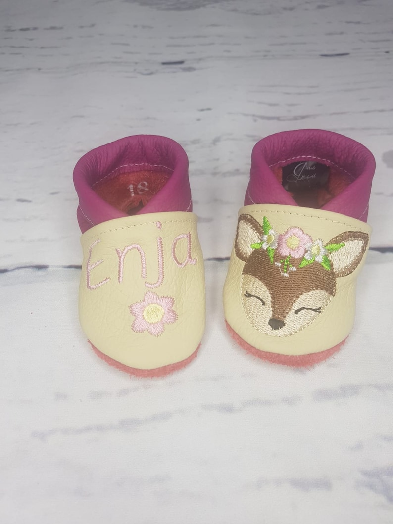 May include: A pair of cream and magenta leather baby shoes. One shoe features the embroidered name Enja in pink with a small flower, while the other displays an embroidered deer face with a floral crown. The interior of the left shoe shows size 18.
