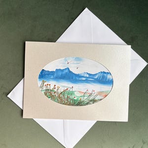 Op de afbeelding: Een wenskaart met een ovaal aquarel schilderij van een kustlandschap. De schildering toont blauwe bergen, een blauwe zee en groene en bruine vegetatie. De kaart zit in een lichtbeige lijst en wordt geleverd met een witte envelop.
