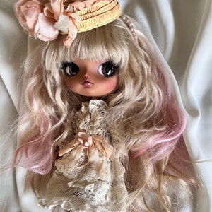 Könnte beinhalten: Eine Puppe mit langen blonden und pinken Haaren, die ein weißes Spitzenkleid und einen Strohhut mit einer rosa Blume trägt.