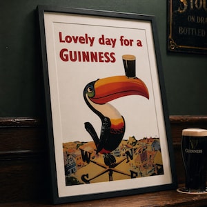 May include: Framed vintage Guinness advertisement featuring a toucan with a pint of Guinness on its beak. The poster reads "Lovely day for a Guinness" in red text. A pint of Guinness sits on a coaster to the right.