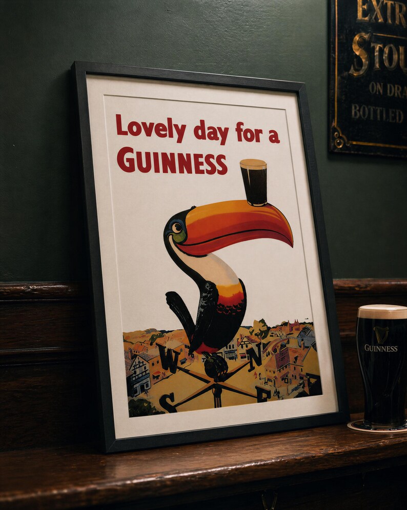 May include: Framed vintage Guinness advertisement featuring a toucan with a pint of Guinness on its beak. The poster reads "Lovely day for a Guinness" in red text. A pint of Guinness sits on a coaster to the right.