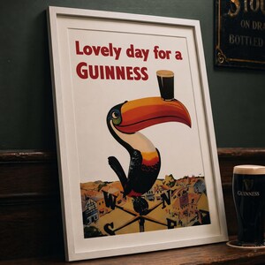 May include: Framed Guinness advertisement featuring a toucan with a pint of Guinness on its beak. The poster reads "Lovely day for a Guinness" in red text. A pint of Guinness sits on a wooden surface next to the framed poster.