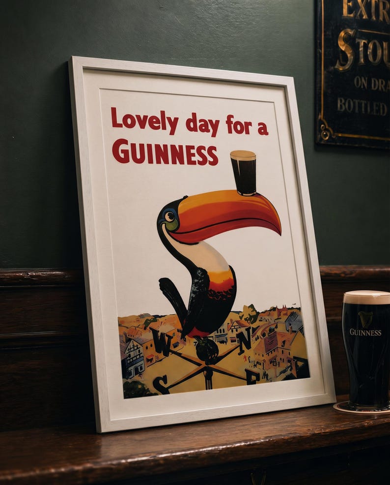 May include: Framed Guinness advertisement featuring a toucan with a pint of Guinness on its beak. The poster reads "Lovely day for a Guinness" in red text. A pint of Guinness sits on a wooden surface next to the framed poster.