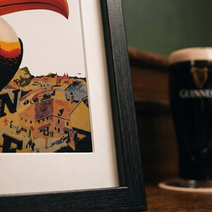 May include: Framed print featuring a toucan with a large orange beak, overlooking a townscape. The artwork is in a black frame, with a white mat. A pint of Guinness stout is in the background, on a wooden surface.