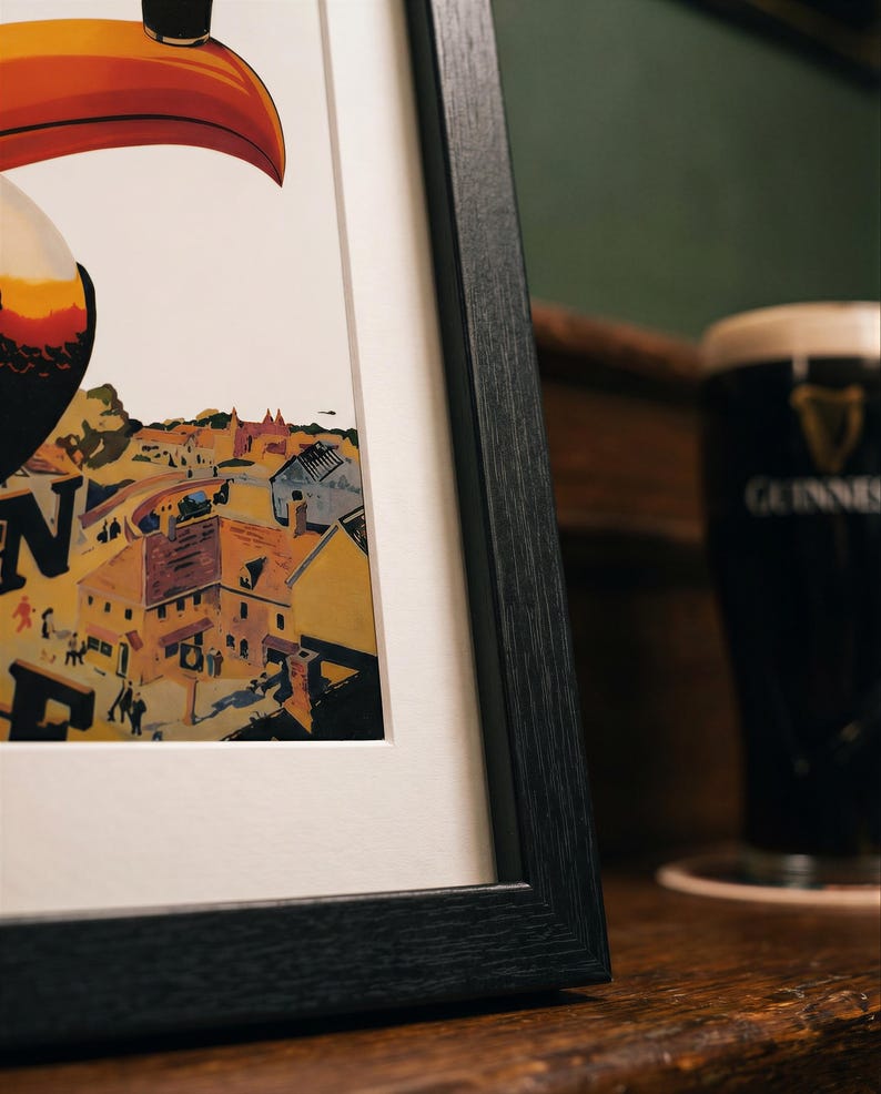 May include: Framed print featuring a toucan with a large orange beak, overlooking a townscape. The artwork is in a black frame, with a white mat. A pint of Guinness stout is in the background, on a wooden surface.