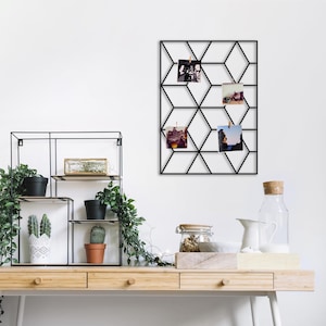 Puede incluir: Panel de exhibición de fotos geométrico negro con cuatro fotos sujetas. El panel está montado en una pared blanca sobre un escritorio de madera. El escritorio tiene una variedad de elementos decorativos, incluidas plantas y un tarro de cristal.