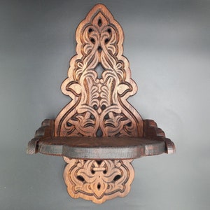 Puede incluir: Una estantería de pared de madera marrón oscuro, tallada intrincadamente, con un diseño ornamentado y simétrico. La estantería presenta un panel trasero decorativo con patrones florales y de volutas, y una pequeña estantería curvada. El diseño es vintage.