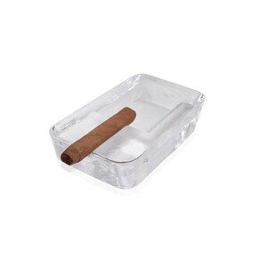 May include: A clear glass ashtray with a brown cigar resting inside. The ashtray is rectangular and has a slightly textured surface.
