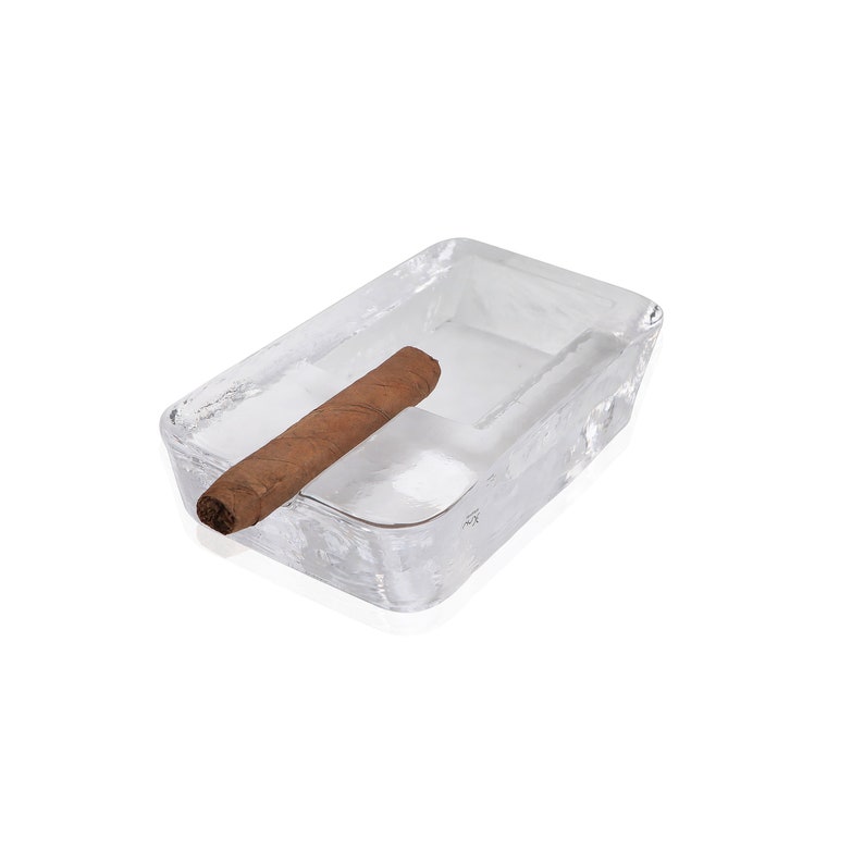May include: A clear glass ashtray with a brown cigar resting inside. The ashtray is rectangular and has a slightly textured surface.