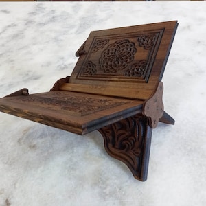May include: A wooden book stand with intricate floral carvings. The stand is made of dark brown wood and features a sloped reading surface and a support that folds out. The stand is designed to hold books or other reading materials.