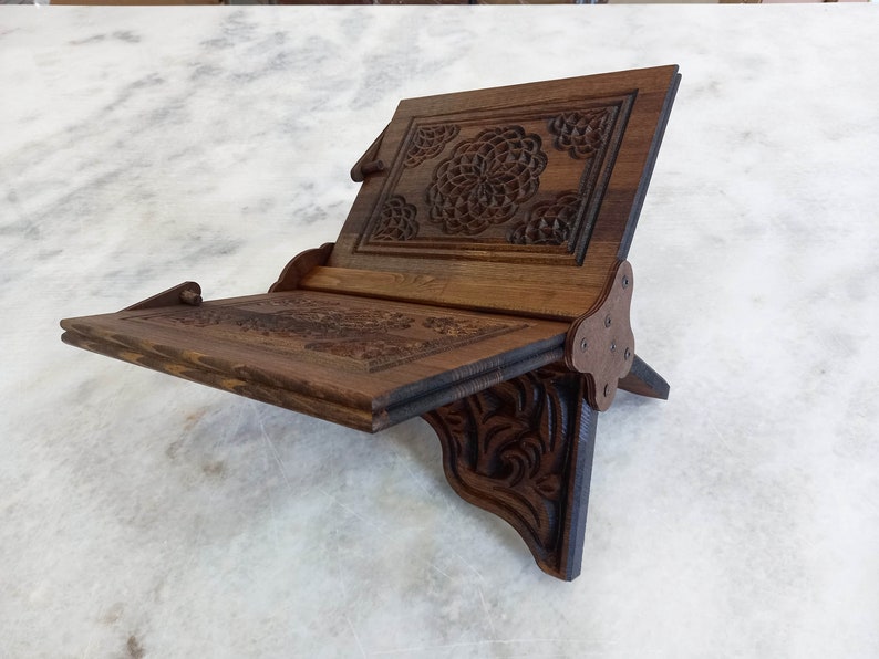 May include: A wooden book stand with intricate floral carvings. The stand is made of dark brown wood and features a sloped reading surface and a support that folds out. The stand is designed to hold books or other reading materials.