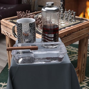 May include: A clear glass ashtray with a brown cigar resting on the edge. The ashtray is on a gray tablecloth, next to a silver and glass tea pot and a silver and black mug. A wooden table with a chessboard is in the background.