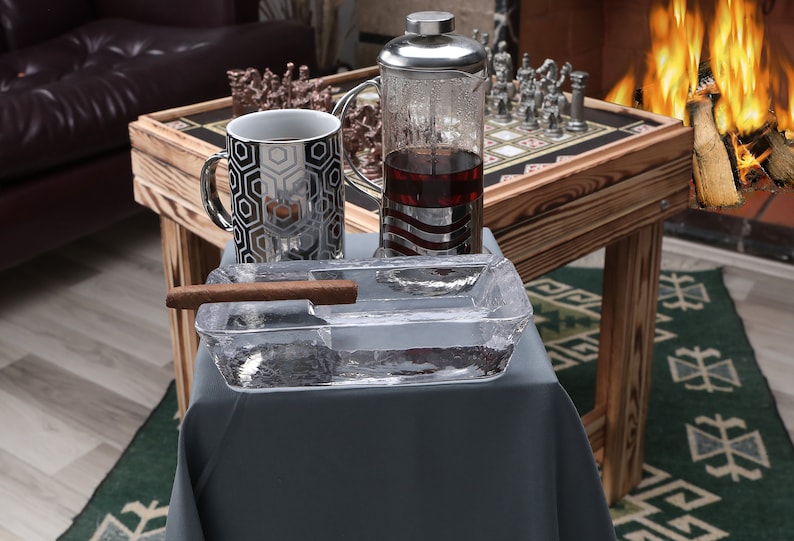 May include: A clear glass ashtray with a brown cigar resting on the edge. The ashtray is on a gray tablecloth, next to a silver and glass tea pot and a silver and black mug. A wooden table with a chessboard is in the background.