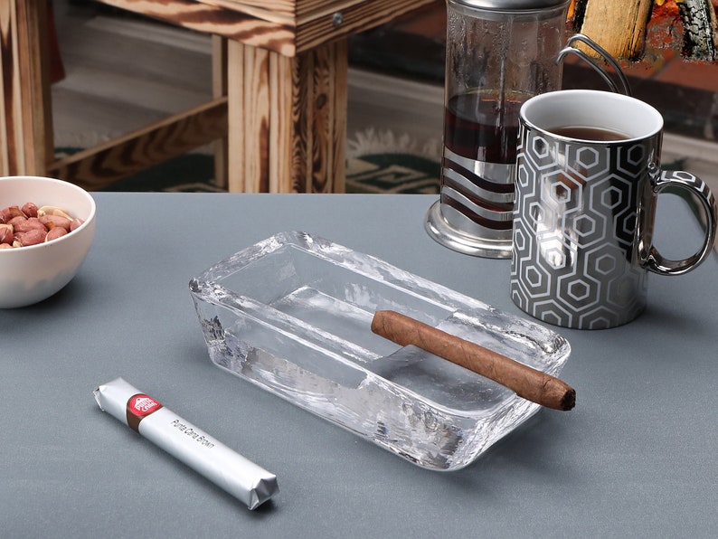 May include: A clear glass ashtray with a cigar resting in the ashtray. The ashtray is on a gray surface. There is a white cup with a geometric pattern and a package of cigars in the background.