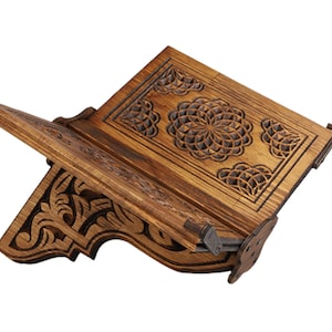May include: A wooden book stand with intricate carved floral designs. The stand is made of light brown wood and features a hinged design for adjustable positioning. The carved details include a central circular pattern and decorative corner elements.
