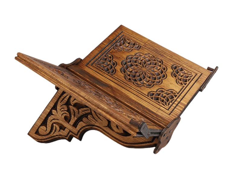 May include: A wooden book stand with intricate carved floral designs. The stand is made of light brown wood and features a hinged design for adjustable positioning. The carved details include a central circular pattern and decorative corner elements.
