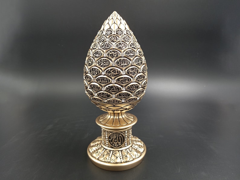 Pode incluir: Um objeto decorativo dourado em forma de pinha, com detalhes intrincados e uma base. O objeto &eacute; coberto com caligrafia &aacute;rabe e pequenos strass transparentes.