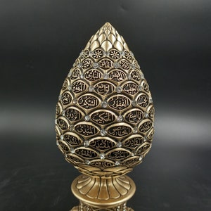Pode incluir: Um objeto decorativo dourado em forma de l&aacute;grima com caligrafia &aacute;rabe preta intrincada e strass transparentes. O objeto est&aacute; sobre uma base dourada.