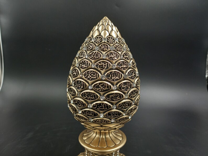 Pode incluir: Um objeto decorativo dourado em forma de l&aacute;grima com caligrafia &aacute;rabe preta intrincada e strass transparentes. O objeto est&aacute; sobre uma base dourada.