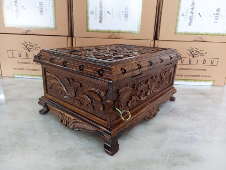 May include: A wooden puzzle box with intricate carvings and a keyhole. The box is dark brown and has a decorative design on the top and sides. The box is sitting on a light brown surface.