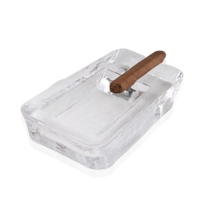 May include: A clear glass ashtray with a rectangular shape and a raised edge. A brown cigar is resting on the edge of the ashtray.