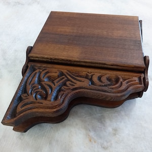 May include: A dark brown wooden shelf with ornate, carved detailing. The shelf has a rectangular top and a decorative, curved front edge with floral and scrollwork patterns. The wood grain is visible, and the shelf appears to be a vintage or antique piece.