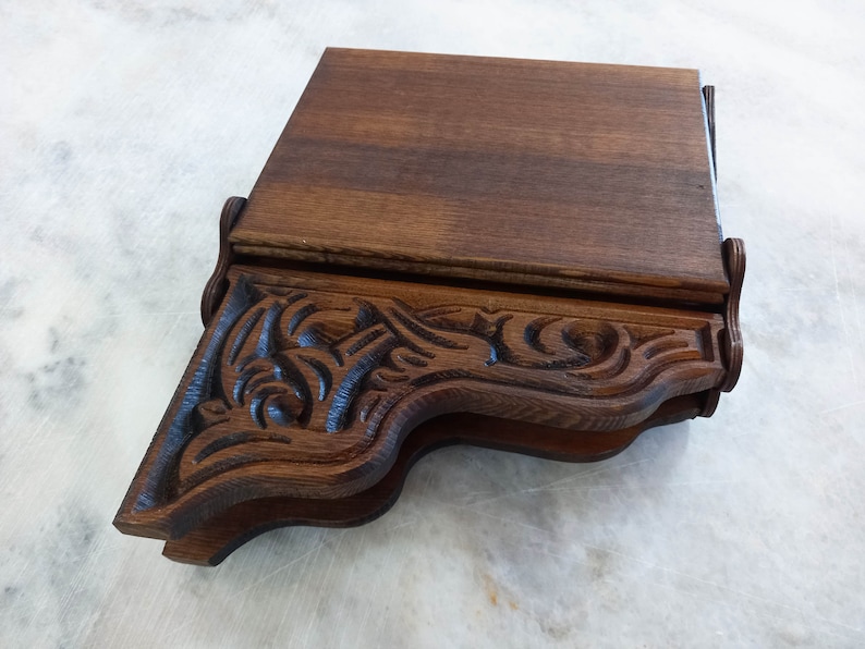 May include: A dark brown wooden shelf with ornate, carved detailing. The shelf has a rectangular top and a decorative, curved front edge with floral and scrollwork patterns. The wood grain is visible, and the shelf appears to be a vintage or antique piece.