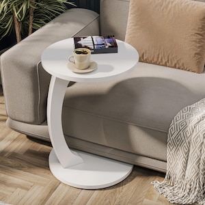 Puede incluir: Mesa auxiliar redonda blanca con base curva. La mesa est&aacute; junto a un sof&aacute; beige y tiene un libro y una taza de caf&eacute; encima.