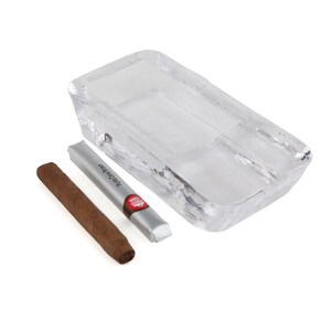 May include: A clear glass ashtray with a rectangular shape and a divided compartment. A brown cigar is resting next to a silver and red package that reads "Premium Cigar".