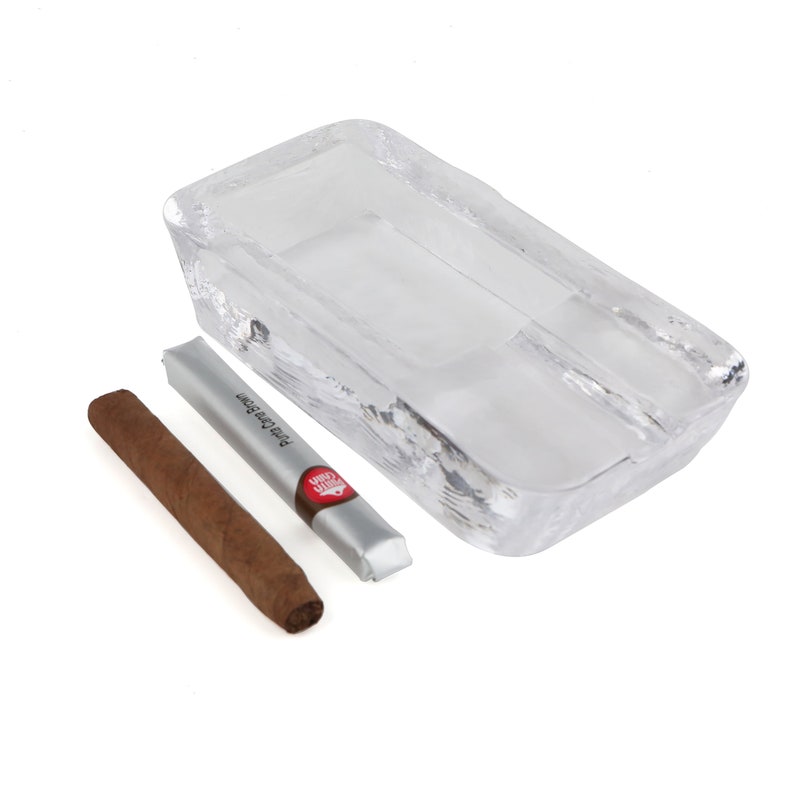 May include: A clear glass ashtray with a rectangular shape and a divided compartment. A brown cigar is resting next to a silver and red package that reads "Premium Cigar".