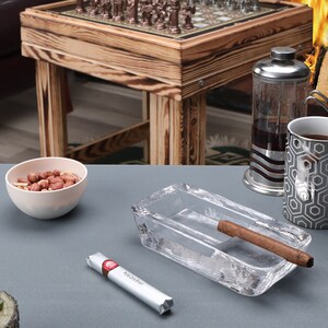 May include: A gray table with a glass ashtray, a cigar, a small bowl of peanuts, a small Christmas tree with a teddy bear, and a silver and black coffee mug.  The mug has a hexagonal pattern.  A silver and glass French press sits behind the mug.