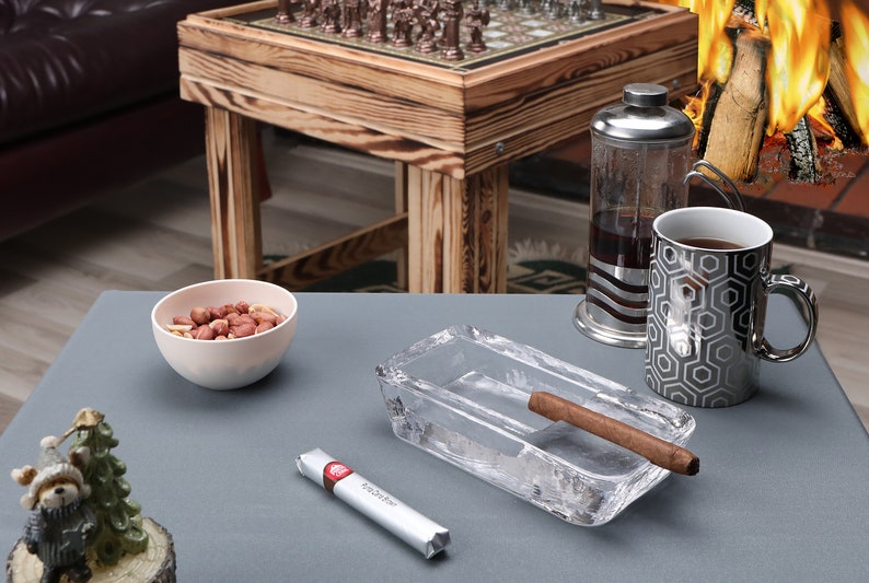 May include: A gray table with a glass ashtray, a cigar, a small bowl of peanuts, a small Christmas tree with a teddy bear, and a silver and black coffee mug.  The mug has a hexagonal pattern.  A silver and glass French press sits behind the mug.