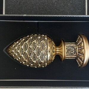 Pode incluir: Um objeto decorativo dourado com detalhes intrincados e uma base em forma de c&uacute;pula. O objeto &eacute; coberto por pequenos padr&otilde;es em forma de diamante em relevo e apresenta caligrafia &aacute;rabe.