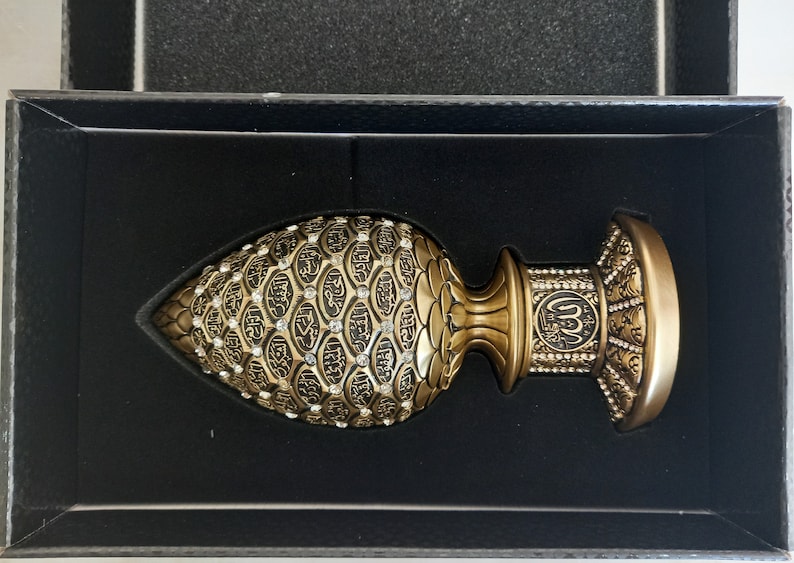 Pode incluir: Um objeto decorativo dourado com detalhes intrincados e uma base em forma de c&uacute;pula. O objeto &eacute; coberto por pequenos padr&otilde;es em forma de diamante em relevo e apresenta caligrafia &aacute;rabe.