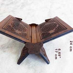 May include: A wooden book stand with intricate floral carvings. The stand is open, displaying the pages, and measures 9.44 inches high and 6.69 inches wide. The dark brown wood has a natural grain, and the stand is supported by decorative legs.