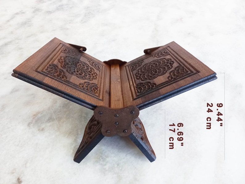 May include: A wooden book stand with intricate floral carvings. The stand is open, displaying the pages, and measures 9.44 inches high and 6.69 inches wide. The dark brown wood has a natural grain, and the stand is supported by decorative legs.
