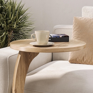 Puede incluir: Una mesa auxiliar de madera clara con una pata curva y una tapa redonda. Una taza de caf&eacute; blanca y un platillo est&aacute;n sobre la mesa, junto con un libro negro con la palabra "ES" visible en la portada.