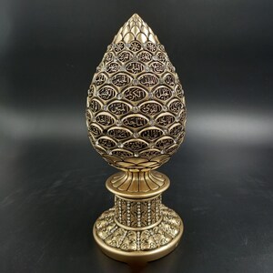 Pode incluir: Um objeto decorativo dourado em forma de l&aacute;grima com caligrafia &aacute;rabe intrincada e strass. O objeto est&aacute; sobre uma base dourada com um design decorativo.