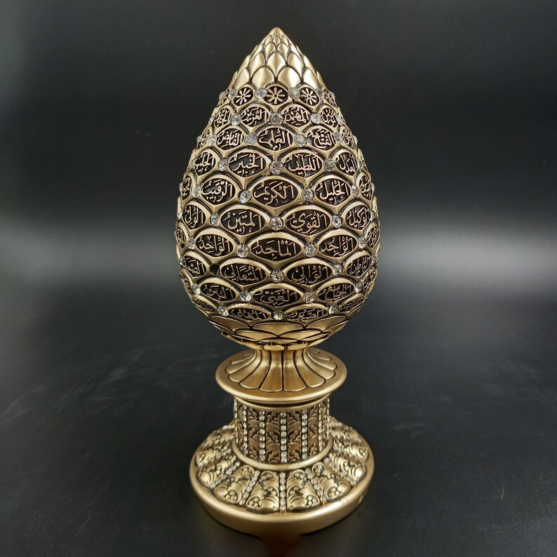 Pode incluir: Um objeto decorativo dourado em forma de l&aacute;grima com caligrafia &aacute;rabe intrincada e strass. O objeto est&aacute; sobre uma base dourada com um design decorativo.