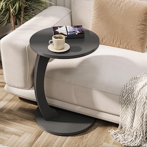 Puede incluir: Una mesa auxiliar de color gris oscuro en forma de C con una tapa redonda. La mesa est&aacute; junto a un sof&aacute; blanco con un coj&iacute;n de tiro beige y una manta. Una taza de caf&eacute; y un libro est&aacute;n sobre la mesa.
