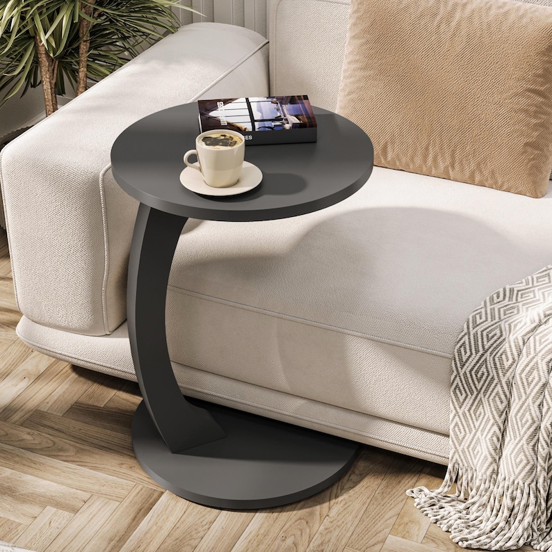Puede incluir: Una mesa auxiliar de color gris oscuro en forma de C con una tapa redonda. La mesa est&aacute; junto a un sof&aacute; blanco con un coj&iacute;n de tiro beige y una manta. Una taza de caf&eacute; y un libro est&aacute;n sobre la mesa.
