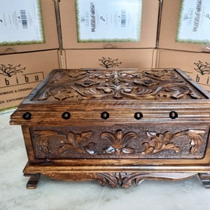 May include: A wooden box with intricate carvings of floral designs. The box has a dark brown stain and is decorated with metal accents. The box is labeled "tubibu" and "PUZZLE BOX".