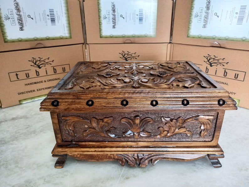May include: A wooden box with intricate carvings of floral designs. The box has a dark brown stain and is decorated with metal accents. The box is labeled "tubibu" and "PUZZLE BOX".