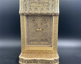 Pode incluir: Uma peça decorativa dourada que retrata uma porta detalhada com padrões intrincados e caligrafia árabe. A peça tem uma moldura semelhante a tijolos e uma pequena base de plataforma, com detalhes e enfeites ornamentados.
