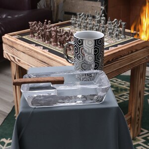 May include: A wooden chessboard with silver and bronze chess pieces, a black and white geometric mug, and a clear glass ashtray with a brown cigar on a wooden table.