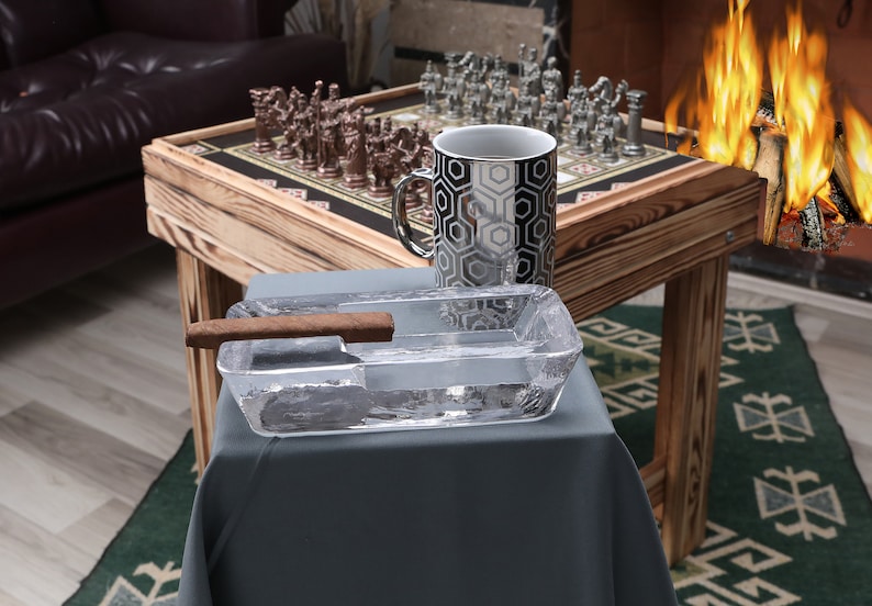 May include: A wooden chessboard with silver and bronze chess pieces, a black and white geometric mug, and a clear glass ashtray with a brown cigar on a wooden table.