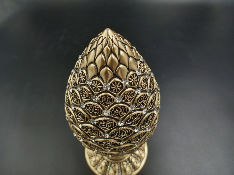 Pode incluir: Um objeto decorativo dourado em forma de pinha, com detalhes intrincados e strass transparentes. O objeto tem uma base com um design semelhante.