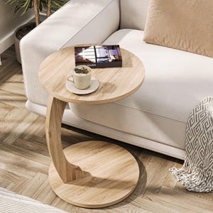 Puede incluir: Una mesa auxiliar de madera clara con una base curva y una tapa redonda. La mesa est&aacute; junto a un sof&aacute; blanco y tiene un libro y una taza de caf&eacute; encima.