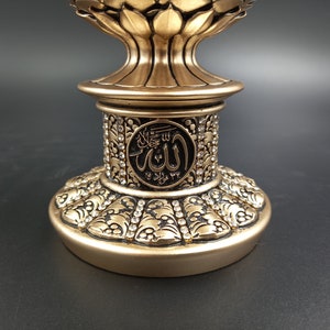 Pode incluir: Um suporte decorativo dourado com detalhes intrincados e uma parte superior redonda. O suporte apresenta um c&iacute;rculo preto com a palavra &aacute;rabe "Allah" em letras brancas.