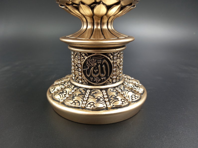Pode incluir: Um suporte decorativo dourado com detalhes intrincados e uma parte superior redonda. O suporte apresenta um c&iacute;rculo preto com a palavra &aacute;rabe "Allah" em letras brancas.